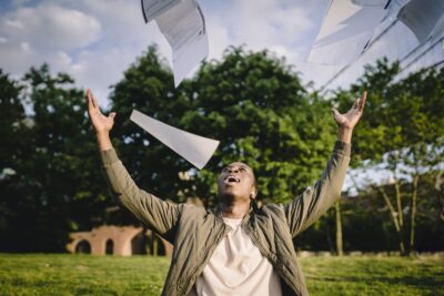 Cheerful young African American male student in casual clothes throwing college papers up in air while having fun in green park after end of exams