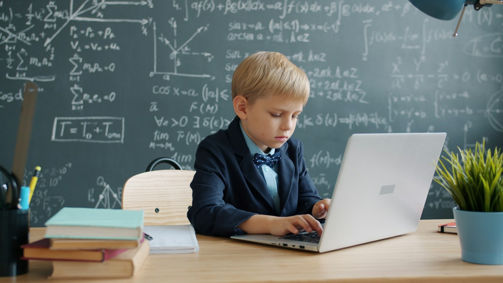 Young boy in suit using laptop at desk with blackboard